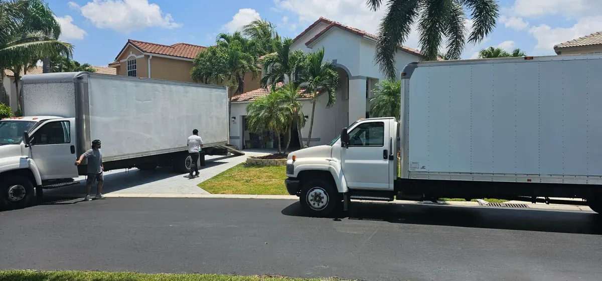 Two white moving trucks and crew members outside a palm-lined home