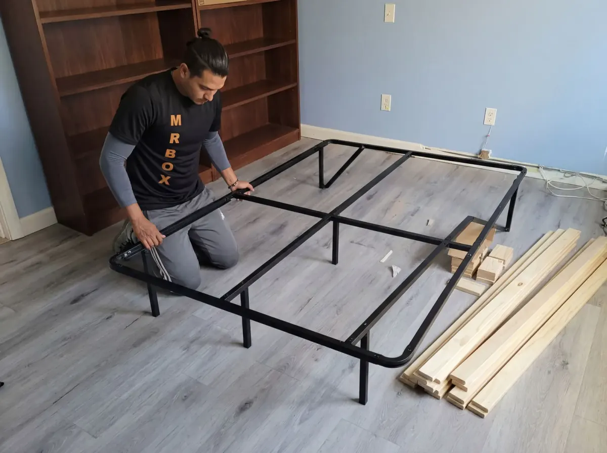 A Mr. Box mover working on a metal bed frame beside stacked wooden slats